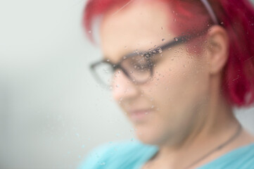 Frau mit roten Haaren und Brille, Nahaufnahme durch Fensterscheibe mit Wassertropfen, unscharfer Hintergrund