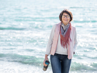 Smiling woman walking on seaside with metal travel mug. Vacation on ocean coast. Turquoise water...