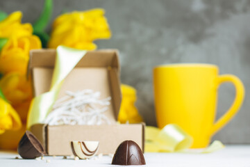 Chocolate candies in craft box, cup and bouquet of yellow tulips on white wooden surface on gray background. Copy space