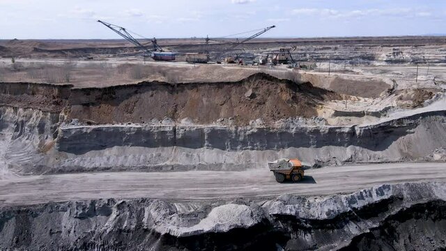 A large dump truck pulls coal soil through the open pit against the backdrop of heavy mining equipment. Coal mine. Side view. Coal transshipment.