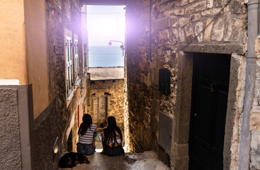 Corniglia, Liguria, Italy. June 2020. Conceptual image of friendship of two young women from behind and a dog sitting next to them
