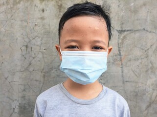 An asian boy wear medical mask in front of  cement wall background. Indonesian boy smile while wear...