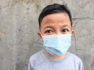 An Asian boy wear medical mask with angry expression in front of  cement wall background. Indonesian boy wear face mask due to corona virus or covid19 pandemic. Avoid covid 19 virus spreading