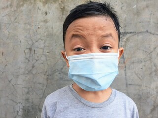 An Asian boy wear medical mask with angry expression in front of  cement wall background. Indonesian boy wear face mask due to corona virus or covid19 pandemic. Avoid covid 19 virus spreading