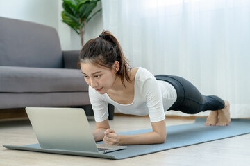 Fototapeta premium Fit, sporty asian young beautiful woman, girl doing yoga planking, watching trainer online on laptop computer, training in living room. Workout fitness exercise person, sporty in sportswear at home.