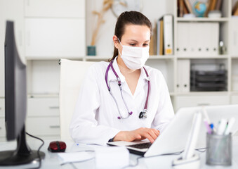 Young female doctor in a protective mask, working in the therapeutic department of the clinic, sits at a desktop in front ..of a laptop in the office