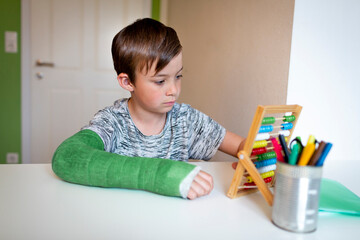 cool boy with green arm cast sits in his room and learns with slide rule with colorful balls made of wood during homeschooling