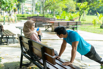 a woman wearing a hijab sportswear drinking with a drinking water bottle while sitting resting when with a male friend doing push up movements on a park bench