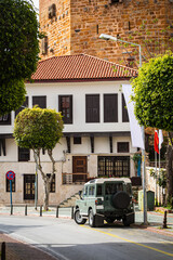 A narrow street along a cafe and a motor road, old jeep  with a paved pathway and an avenue of green trimmed trees against the backdrop of an old fortress made of crane bricks and