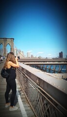 a woman taking photos of the Dumbo neighborhood in Brooklyn, New York, from the Brooklyn Bridge and with the Manhattan skyline in the background
