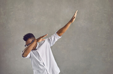 Happy handsome young black man with Afro hairstyle dancing dub against gray stone street wall. Funky African American guy in white shirt doing popular dab dance arm move on concrete urban background - obrazy, fototapety, plakaty
