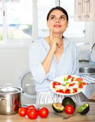 Young woman eating fresh vegetable salad with cheese in kitchen