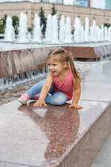 happy caucasian child girl crawling on the side of the fountain in the city park