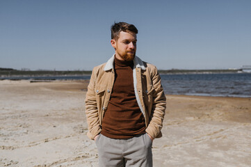 A young man with a beard walks by the river.