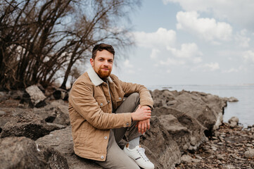 A young man with a beard is sitting on the rocks by the river.