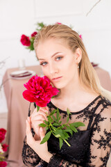 An attractive young girl with long blond hair and evening make-up in a black long evening dress, surrounded by live burgundy peonies and a festively set table for dinner. Spring mood. 
