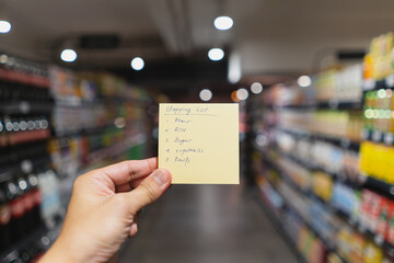 Hand holding a yellow note with hand writing shopping list in blur supermarket aisle background