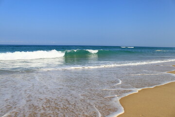 Fototapeta premium Une vague avec la mousse blanche avance vers la plage de sable jaune. L´océan est bleu vert et le ciel est bleu sans aucun nuage.