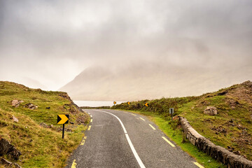 Small narrow asphalt road in a mountains by a lake, low clouds cover mountains peaks, fog...
