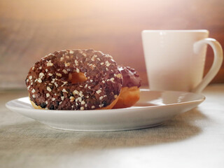 Doughnut with chocolate icing on a white plate and white tea cup, sun light flare in the background. Breakfast concept. Pastry product. Delicious and unhealthy choice of desert