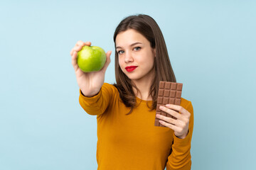 Teenager girl isolated on blue background taking a chocolate tablet in one hand and an apple in the...