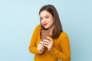 Teenager girl isolated on blue background taking a chocolate tablet and happy