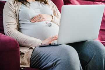 Unrecognizable young pregnant woman working remotely sitting on sofa at home with laptop touching her belly with her hand - Businesswoman working during maternity