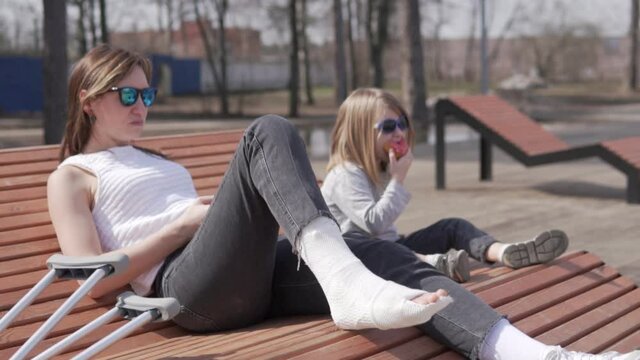 A woman with plaster on her leg sits on a bench in the park with a small daughter, the family has a picnic, Focus on the plastered leg, mother and child blurred.