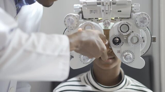 Optician doing optometry eye exam for black african american teen girl patient. Male optometrist with phoropter while examining patient.