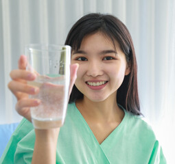 Close up of happy young inpatient woman  who is getting well from her ailment smiling on to camera and holding a glass of water in hand as a foreground.