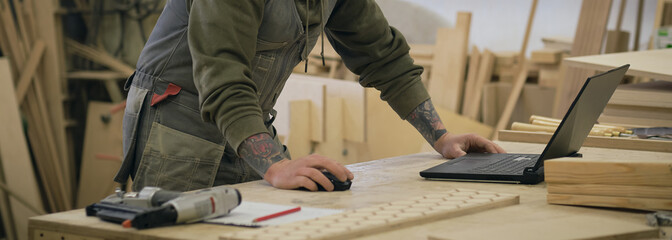 Concentrated adult craftsman checking information on laptop while working in carpentry workshop....