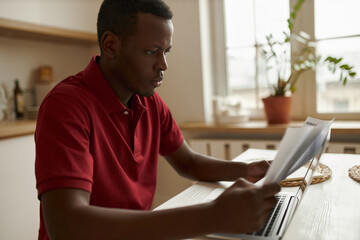 Successful young African entrepreneur studying financial papers with attentive concentrated look, sitting at kitchen table using portable computer. Concentrated focused black man paying bills online