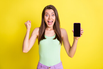 Photo of crazy astonished lady show phone screen empty space raise fist wear green top isolated yellow color background