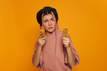 Studio shot of fashionable cute young Caucasian woman wearing synthetic box braids staring at camera with angry look frowning eyebrows, aiming two bananas at you as if shooting guns, playing