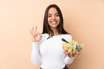 Young brunette girl holding a salad over isolated background showing an ok sign with fingers