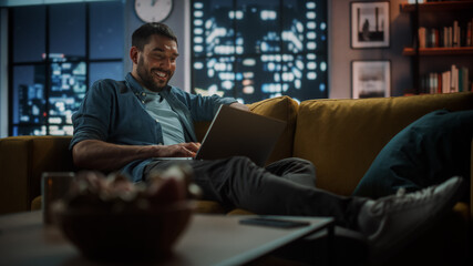 Handsome Caucasian Man Working on Laptop Computer while Resting on a Sofa in Dark Cozy Living Room in the Evening. Freelancer Working From Home. Browsing Internet, Using Social Networks, Having Fun.