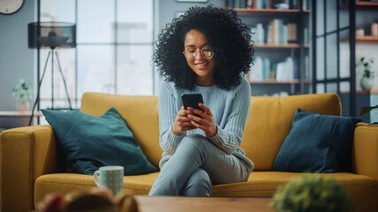 Portrait of a Beautiful Authentic Latina Female in a Stylish Cozy Living Room Using Smartphone at Home. She's Browsing the Internet and Checking Videos on Social Networks and Having Fun. - obrazy, fototapety, plakaty