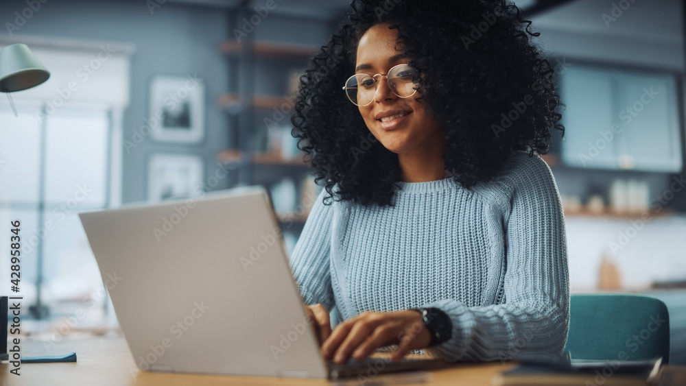Poster beautiful authentic latina female with afro hair sitting at a desk in a cozy living room and using l