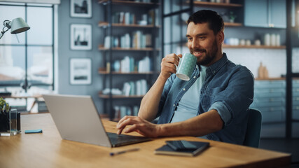 Handsome Caucasian Man Working on Laptop Computer while Sitting on a Sofa Couch in Stylish Cozy Living Room. Freelancer Working From Home. Browsing Internet, Drinking Coffee from a Mug, Having Fun.