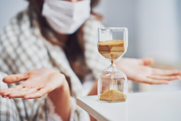 Adult woman in mask feeling sick at home with hourglass Covid-19 concept