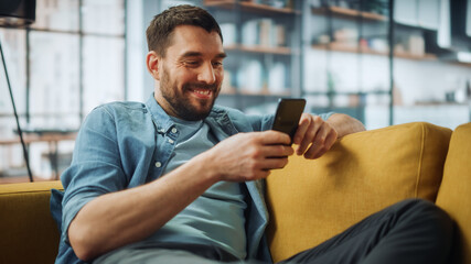 Happy Handsome Caucasian Man Using Smartphone in Cozy Living Room at Home. Man Resting on Comfortable Sofa. He's Browsing the Internet and Checking Videos on Social Networks and Having Fun.