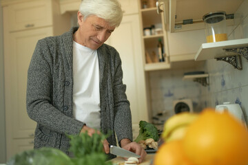 Nutrition, health and aging concept. Attractive mature man with gray hair standing at kitchen counter surrounded with fresh organic ingredients, slicing mushrooms for soup using sharp knife