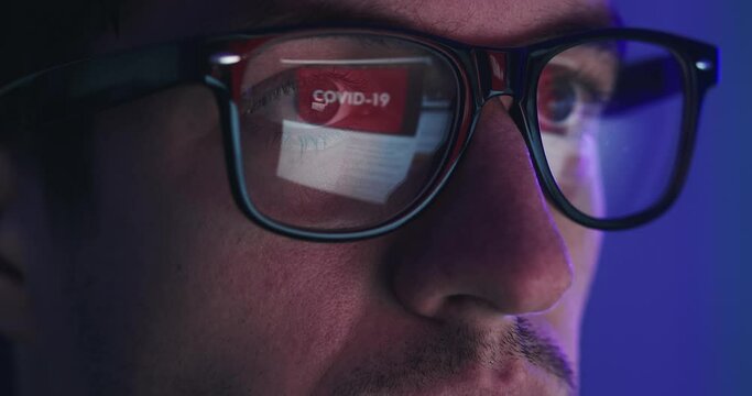 Close-up portrait of man in eyeglasses late at night scrolling news abous coronavirus in front of laptop