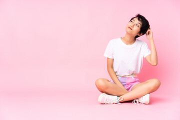 Young Vietnamese woman with short hair sitting on the floor over isolated pink background having doubts and with confuse face expression