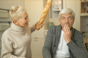 Angry mature wife posing in kitchen with french bread in hand going to hit her scared husband, having dispute, sorting out relationships. Funny senior couple fooling around, playing with food
