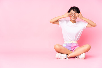Young Vietnamese woman with short hair sitting on the floor over isolated pink background covering eyes by hands