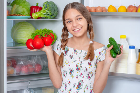 Smiling young teen girl holding fresh red tomatoes and green cucumbers while standing near open fridge in kitchen at home. Portrait of child choosing food in refrigerator full of healthy products