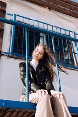 Smiling young woman in black leather jacket sitting on stairs of watchtower in front of blue railings. Having fun outside. Urban style and street fashion. Girl with closed eyes in casual outfit