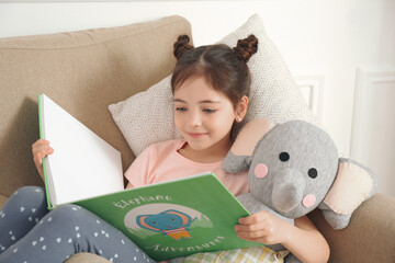 Little girl with toy elephant reading book in armchair at home