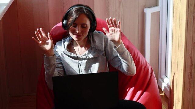 Happy Young European Woman With Short Hair In Headphones In Grey Clothes Sitting On Red Bag Chair At Balcony Dancing And Singing Using Laptop Computer. Joyful Student Studying At Home.